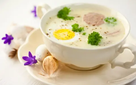 Aromaticzny barszcz biały na żeberkach z jajkiem i kiełbasą, ozdobiony natką pietruszki. Na talerzyku wielkanocne jajka i fioletowe kwiaty.
