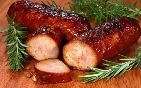 Soczysta kiełbasa pieczona w piekarniku, skropiona błyszczącym sosem, z gałązkami rozmarynu i ziarnami pieprzu.