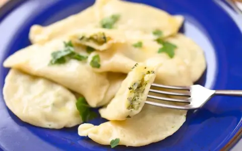 Pyszne pierogi ze szpinakiem i serem, udekorowane natką pietruszki, na niebieskim talerzu. Jedna sztuka jest nadziana widoczna na widelcu.