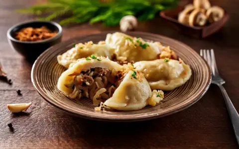 Talerz pełen pysznych, domowych pierogów z kapustą i grzybami, posypanych szczypiorkiem. Obok czosnek i pieprz.