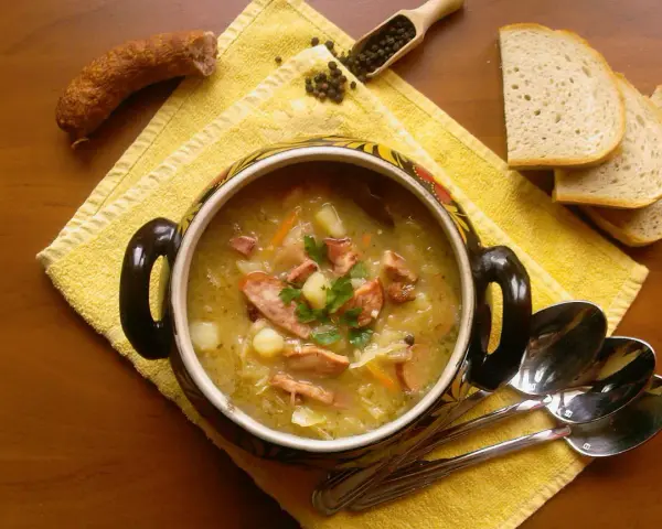 Aromaticzny kapuśniak z kiszonej kapusty, podany w ozdobnym garnku, z kiełbasą, ziemniakami i pieczywem.