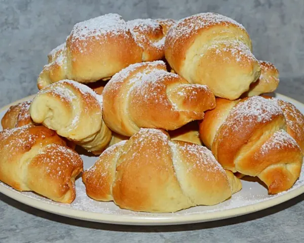 Stos świeżo upieczonych rogalików z czekoladą, posypanych cukrem pudrem, na talerzu.