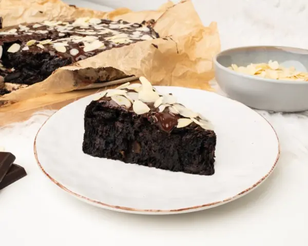 Wilgotny brownie z buraka, polany czekoladą i posypany płatkami migdałów, na talerzu.