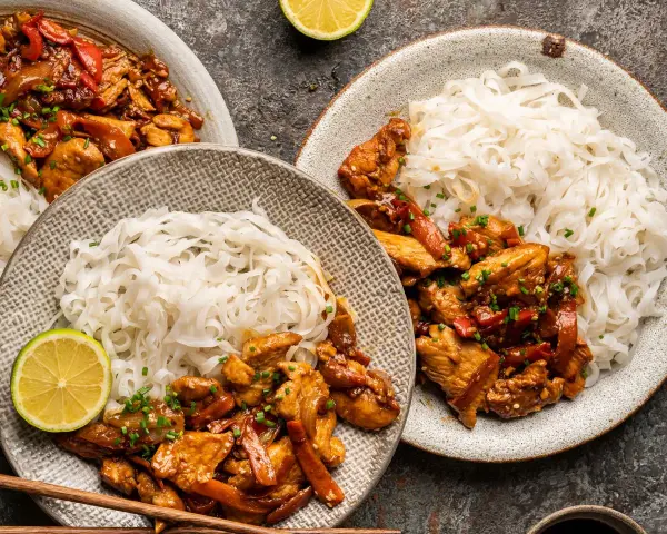 Dwa talerze z kurczakiem teriyaki i makaronem ryżowym, udekorowane plasterkami limonki i szczypiorkiem.