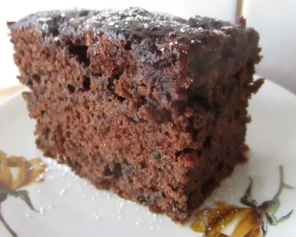 Wilgotne ciasto czekoladowe z dżemem porzeczkowym, posypane cukrem pudrem, na talerzyku z motywem kwiatowym.