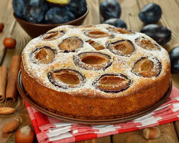 Pyszne ciasto ze śliwkami, posypane cukrem pudrem, na drewnianym blacie. Obok leżą orzechy, migdały i laski cynamonu.