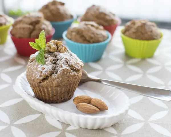 Pyszne muffinki bananowe fit, posypane cukrem pudrem, z orzechami i miętą. Obok migdały i łyżeczka.