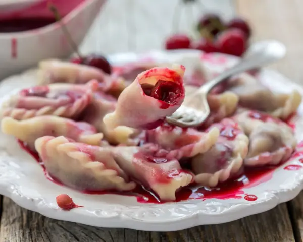 Pyszne pierogi z wiśniami, polane słodkim sosem, kuszą swoim wyglądem. Jedna sztuka jest podniesiona widelcem, ukazując soczyste nadzienie.