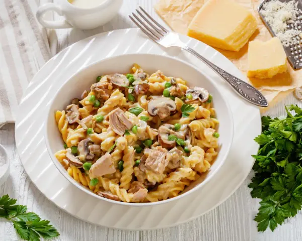 Makaron z tuńczykiem z puszki, groszkiem i pieczarkami w kremowym sosie, posypany natką pietruszki. Obok kawałki parmezanu i tarka.