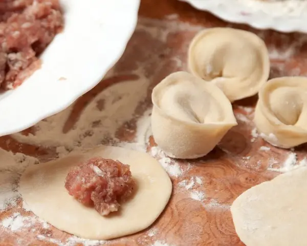 Przygotowanie pierogów z mięsem mielonym surowym. Na drewnianej desce widać surowe ciasto, farsz i gotowe pierożki.