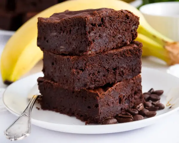 Stos puszystych brownie z bananami, ozdobiony ziarnami kawy, na talerzu z bananem w tle.