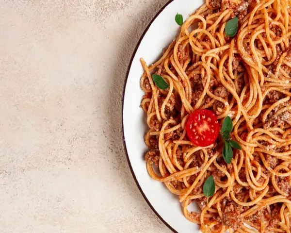 Talerz spaghetti bolognese z pomidorkiem i bazylią na jasnym tle.