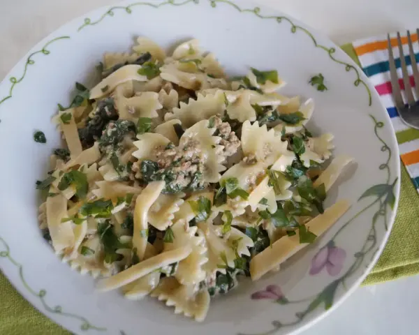 Pyszny makaron z pesto i tuńczykiem, posypany świeżą natką pietruszki, podany na talerzu z ozdobnym brzegiem.