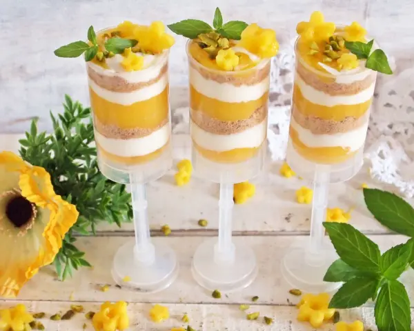 Warstwowy deser z mango w przezroczystych pojemnikach, ozdobiony listkami mięty i pistacjami.