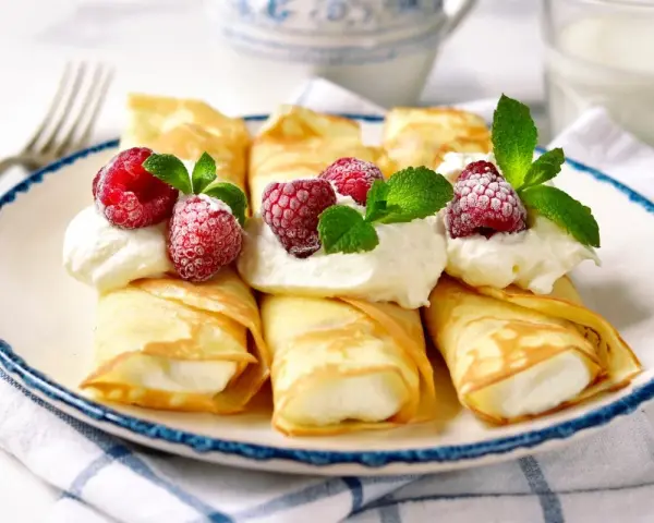 Rolowane naleśniki z mascarpone, udekorowane malinami i miętą, wyglądają apetycznie.