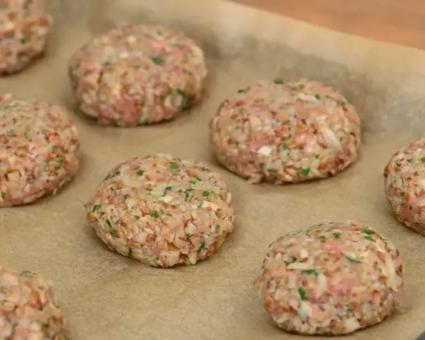 Surowe burgery z piekarnika, przygotowane z mielonego mięsa i kaszy, leżą na papierze do pieczenia.