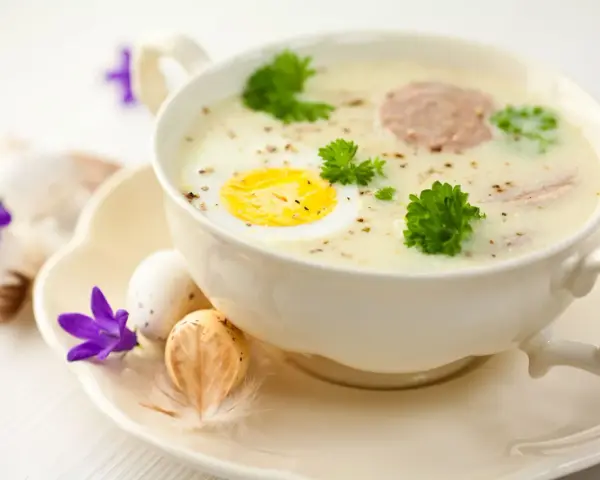 Aromaticzny barszcz biały na żeberkach z jajkiem i kiełbasą, ozdobiony natką pietruszki. Na talerzyku wielkanocne jajka i fioletowe kwiaty.