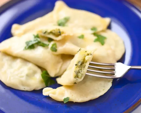 Pyszne pierogi ze szpinakiem i serem, udekorowane natką pietruszki, na niebieskim talerzu. Jedna sztuka jest nadziana widoczna na widelcu.