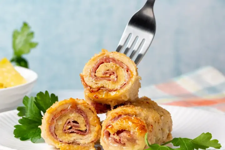 Roladki z piersi kurczaka z szynką i serem, jedna nabita na widelec, na talerzu z natką pietruszki.
