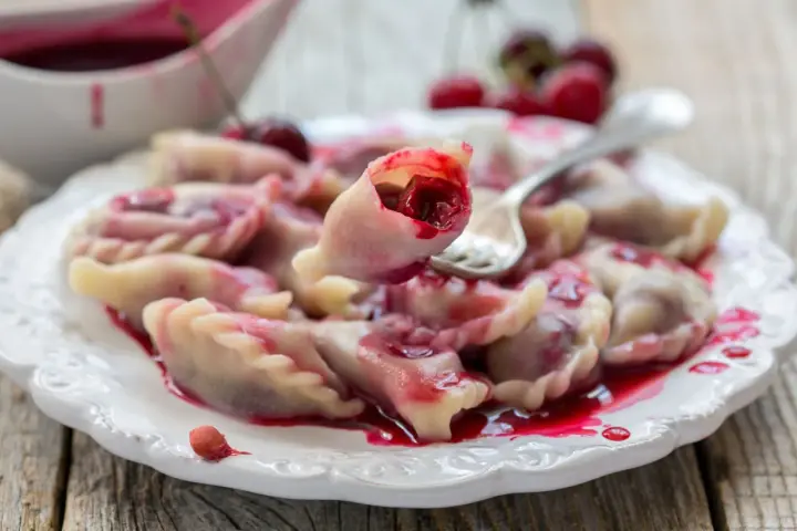 Pyszne pierogi z wiśniami, polane słodkim sosem, kuszą swoim wyglądem. Jedna sztuka jest podniesiona widelcem, ukazując soczyste nadzienie.