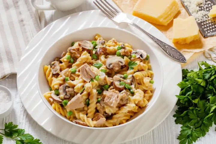 Makaron z tuńczykiem z puszki, groszkiem i pieczarkami w kremowym sosie, posypany natką pietruszki. Obok kawałki parmezanu i tarka.
