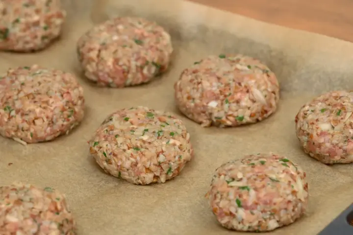 Surowe burgery z piekarnika, przygotowane z mielonego mięsa i kaszy, leżą na papierze do pieczenia.