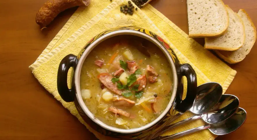 Aromaticzny kapuśniak z kiszonej kapusty, podany w ozdobnym garnku, z kiełbasą, ziemniakami i pieczywem.