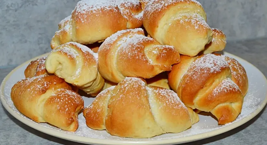 Stos świeżo upieczonych rogalików z czekoladą, posypanych cukrem pudrem, na talerzu.