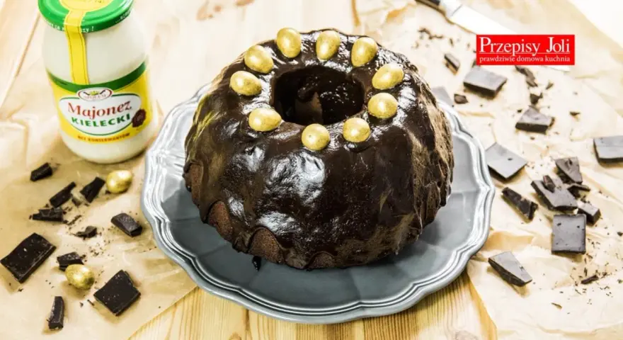 Babka majonezowa z przepisów Joli, polana czekoladą i ozdobiona złotymi jajeczkami. Obok słoik majonezu i kawałki czekolady.