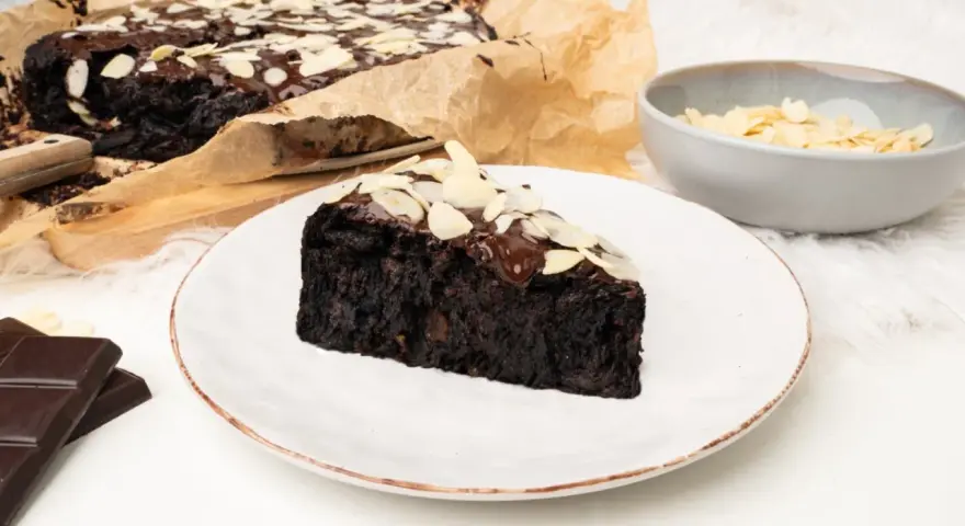 Wilgotny brownie z buraka, polany czekoladą i posypany płatkami migdałów, na talerzu.