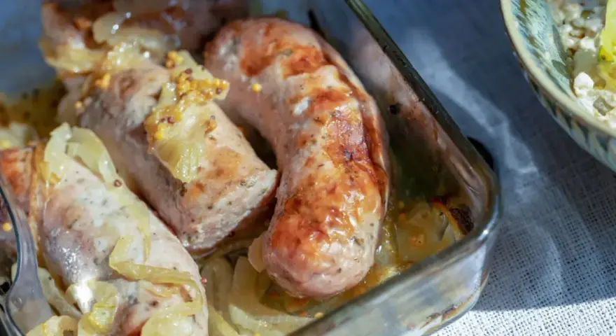 Biała kiełbasa pieczona z cebulką i musztardą w szklanym naczyniu. Apetyczny zapach unosi się w powietrzu.