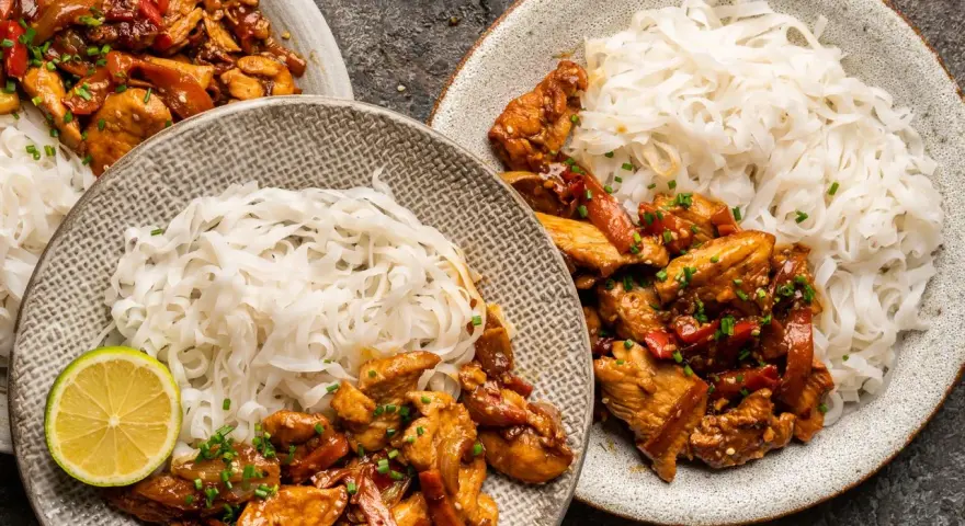 Dwa talerze z kurczakiem teriyaki i makaronem ryżowym, udekorowane plasterkami limonki i szczypiorkiem.