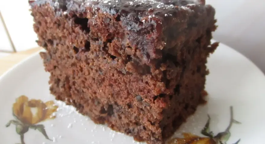 Wilgotne ciasto czekoladowe z dżemem porzeczkowym, posypane cukrem pudrem, na talerzyku z motywem kwiatowym.