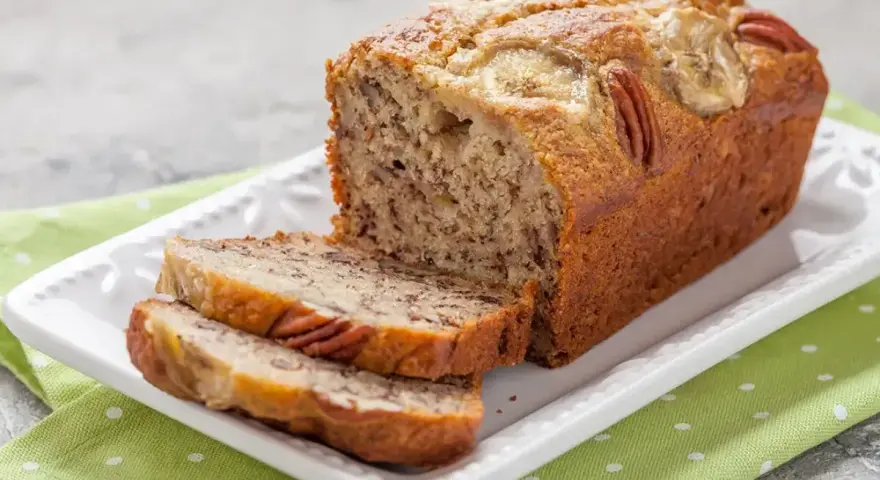 Wilgotny chlebek bananowy z orzechami pekan i plasterkami banana na wierzchu, pokrojony na kromki, podany na białej paterze z zieloną serwetką w groszki.