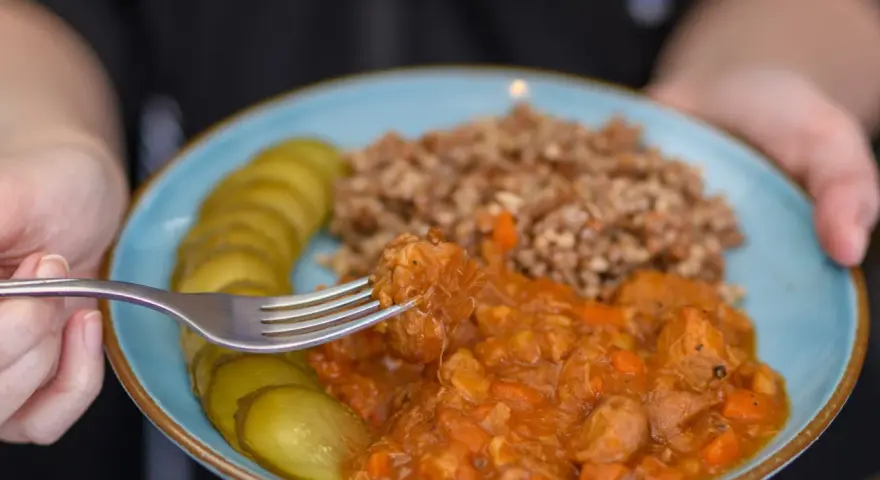 Pyszny gulasz z kaszą gryczaną i ogórkami kiszonymi na talerzu. Widok z bliska na kawałek mięsa nabierany widelcem.
