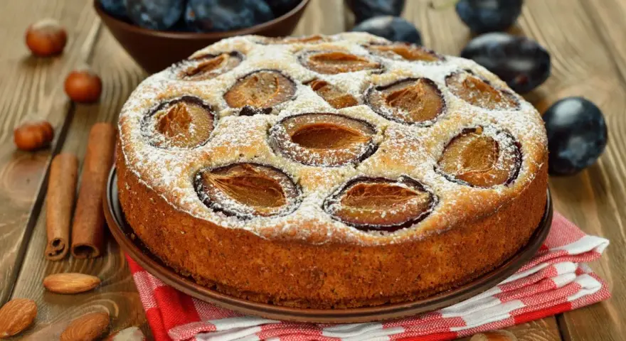 Pyszne ciasto ze śliwkami, posypane cukrem pudrem, na drewnianym blacie. Obok leżą orzechy, migdały i laski cynamonu.