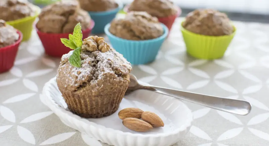 Pyszne muffinki bananowe fit, posypane cukrem pudrem, z orzechami i miętą. Obok migdały i łyżeczka.