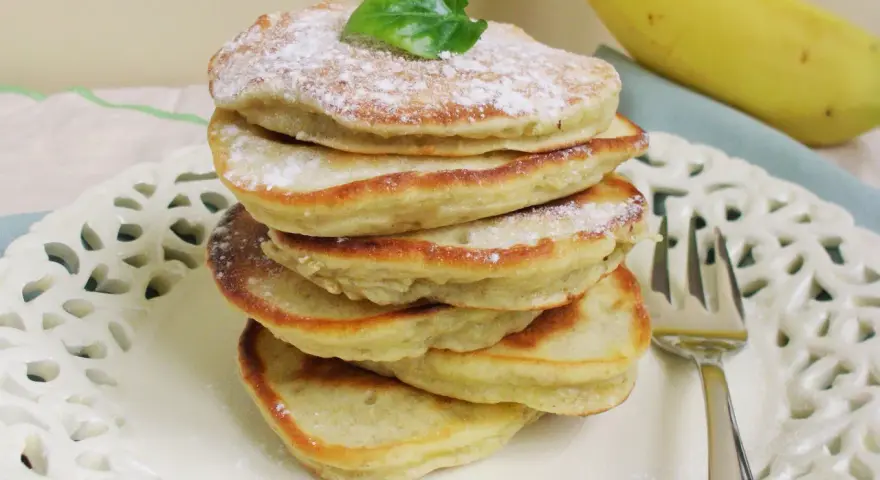 Stosik puszystych placuszków bananowych bez mleka, posypanych cukrem pudrem, z bananem w tle.