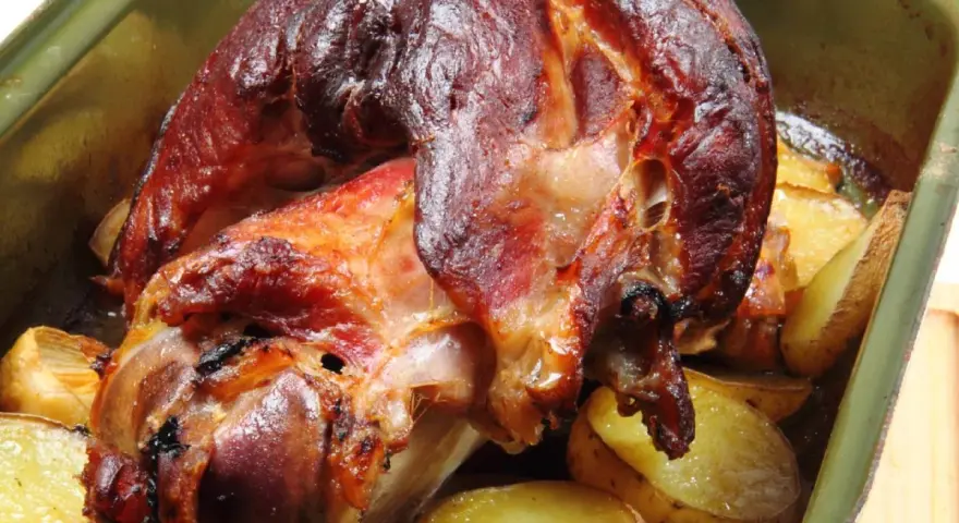 Pieczona golonka w zielonej brytfance, otoczona ziemniakami. Idealna marynata do golonki sprawiła, że skórka jest chrupiąca.