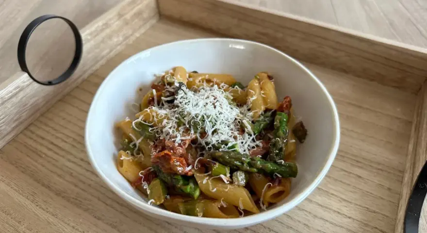 Pyszny makaron ze szparagami i pomidorami, posypany startym parmezanem, podany w białej misce na drewnianej tacy.
