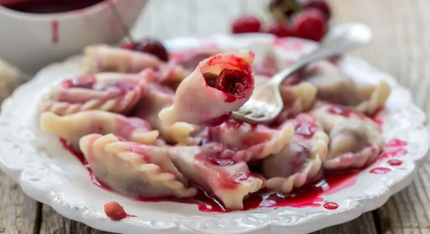 Pyszne pierogi z wiśniami, polane słodkim sosem, kuszą swoim wyglądem. Jedna sztuka jest podniesiona widelcem, ukazując soczyste nadzienie.