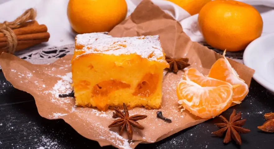 Puszyste ciasto z mandarynkami, posypane cukrem pudrem, obok świeże cząstki owoców i gwiazdki anyżu.