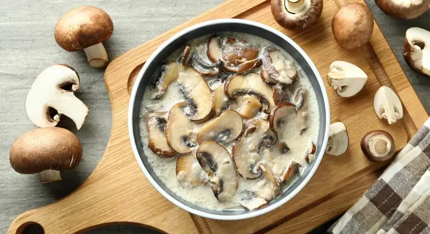 Pyszny sos grzybowy z mrożonych grzybów, podany w misce na drewnianej desce, otoczony świeżymi pieczarkami.