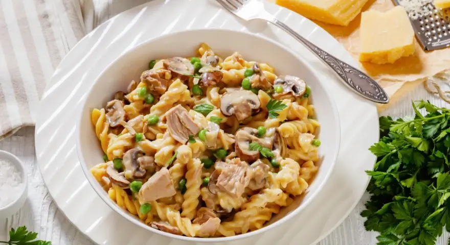 Makaron z tuńczykiem z puszki, groszkiem i pieczarkami w kremowym sosie, posypany natką pietruszki. Obok kawałki parmezanu i tarka.