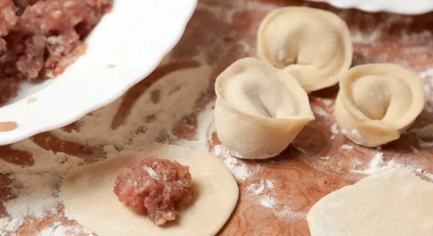 Przygotowanie pierogów z mięsem mielonym surowym. Na drewnianej desce widać surowe ciasto, farsz i gotowe pierożki.