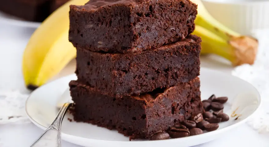 Stos puszystych brownie z bananami, ozdobiony ziarnami kawy, na talerzu z bananem w tle.