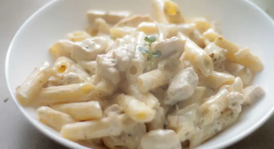 Penne w kremowym sosie serowym z kawałkami kurczaka, udekorowane gałązką ziół. Pyszny makaron serowy z kurczakiem.