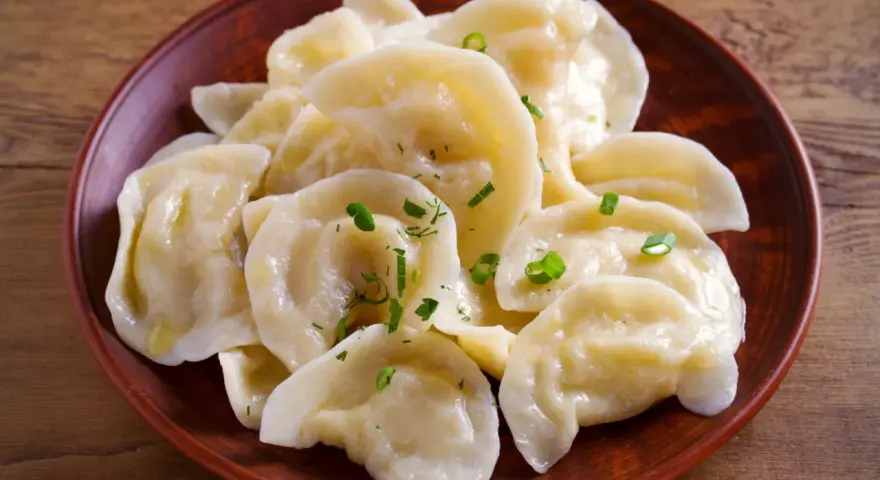 Pyszne pierogi z tradycyjnym farszem do uszek, posypane szczypiorkiem, na glinianym talerzu.