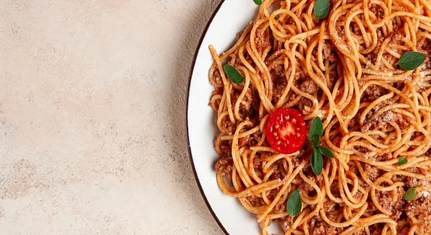 Talerz spaghetti bolognese z pomidorkiem i bazylią na jasnym tle.