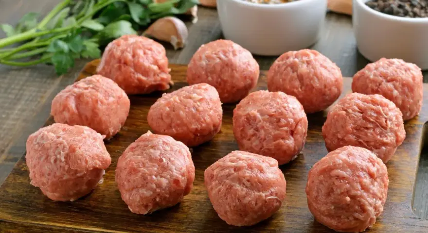 Surowe kotlety mielone uformowane w kulki, gotowe do smażenia. Obok pęczek natki, czosnek i przyprawy.
